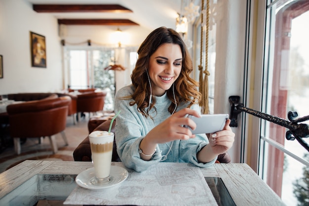 Mulher feliz com cachos no café usando telefone inteligente e sorrindo Foto gratuita