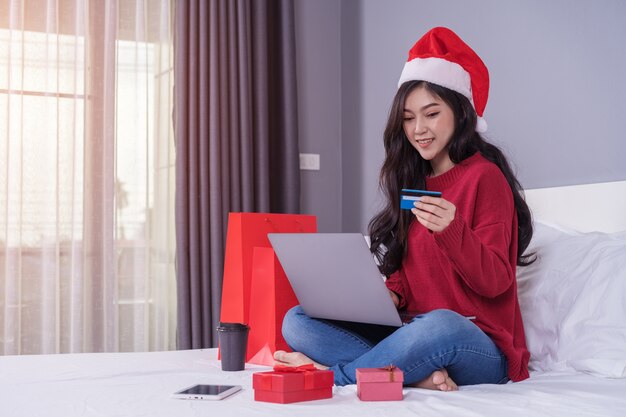 Mulher feliz compras on-line para presente de natal com computador portátil e cartão de crédito na cama Foto Premium