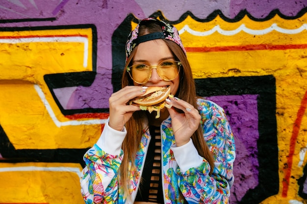 Mulher jovem e atraente, comendo um hambúrguer saboroso. vestida com jaqueta e boné coloridos Foto gratuita