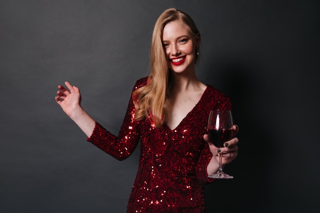 Mulher loira sorridente bebendo vinho tinto. foto de estúdio de menina bonita em vestido de dança em fundo preto. Foto gratuita