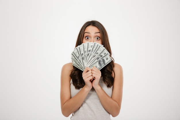 Mulher morena animada, cobrindo o rosto com leque de notas de dólar, expressando choque e surpresa sobre parede branca Foto gratuita