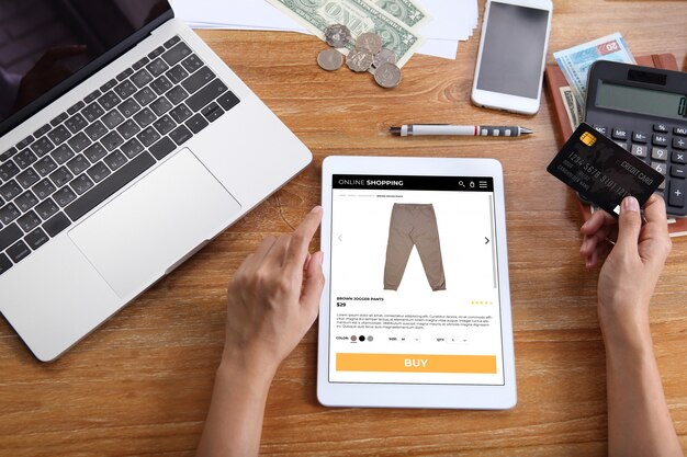 Mulher usando cartão de crédito para comprar calças de jogger marrom no site de comércio eletrônico via tablet com laptop, smartphone e escritório de papelaria na mesa de madeira Foto Premium