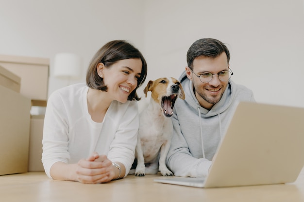 O homem e a mulher sorridente trabalham no computador portátil moderno, boceja de cachorro, compra móveis para o apartamento novo, deita-se no chão na espaçosa sala de luz, tem expressões alegres. conceito de inauguração e reparo Foto Premium