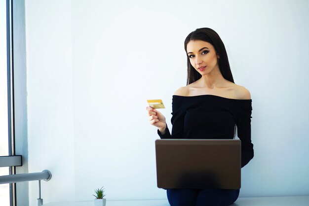 O negócio. mulher de negócios jovem segurando o cartão de crédito nas mãos. senta-se em cima da mesa no escritório big light e trabalha no computador. trabalhe em casa. alta resolução Foto Premium