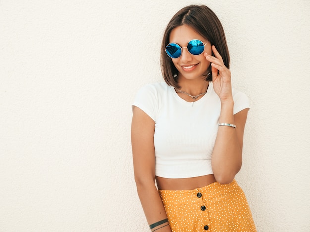 O Retrato Do Modelo Moreno Sorridente Bonito Vestiu Se Com Roupas De Hipster De Verao Menina Na Moda Posando Na Rua Perto Da Parede Branca Mulher Engracada E Positiva Se Divertindo Foto