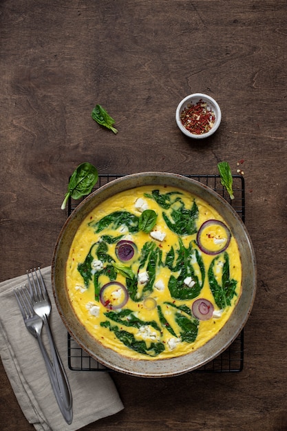 Omelete caseiro, delicioso ou fritada com espinafre, queijo feta, cebola roxa em uma panela de ferro sobre uma mesa rústica. vista do topo Foto Premium