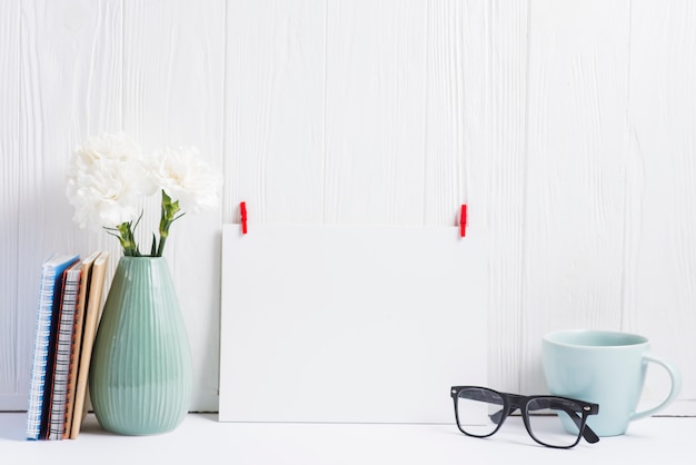 Papel em branco branco com cabide vermelho; óculos; copo; vaso e livros sobre o pano de fundo texturizado de madeira Foto gratuita