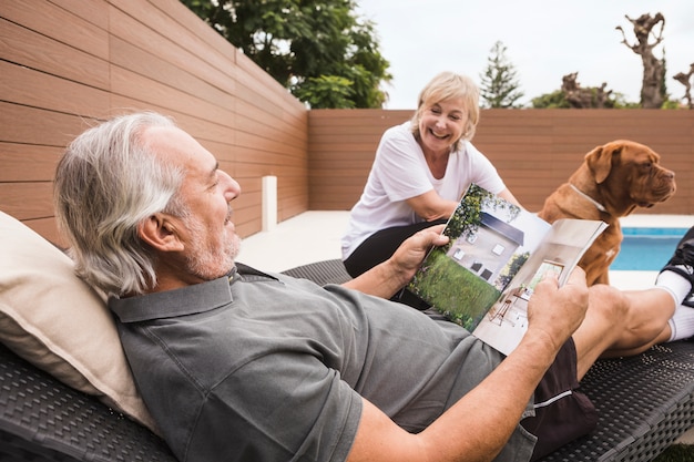 na imagem um homem e uma mulher idosos aproveitam seu tempo após a aposentadoria juntos com um cachorro em um jardim com piscina | Como planejar e como funciona a aposentadoria?