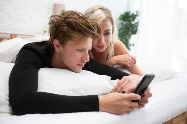 Pares novos com o smartphone que relaxa ao reclinar no noivo que senta-se na cama no quarto em casa. Foto Premium