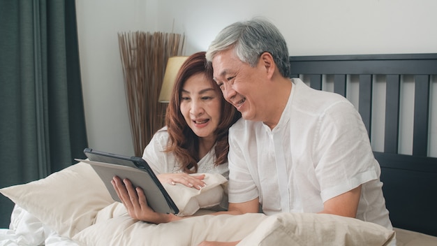 Pares sênior asiáticos usando a tabuleta em casa. as avós chinesas sênior asiáticas, vídeo-chamada que falam com neto da família caçoam ao encontrar-se na cama no quarto em casa no conceito da manhã. Foto gratuita