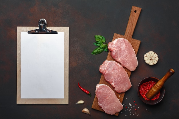 Pedaços de bife de carne de porco crua com manjericão, alho, pimenta, sal e especiaria argamassa e tablet na placa de corte e fundo marrom enferrujado com espaço para seu texto. Foto Premium