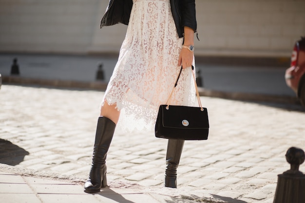 Pernas de uma jovem mulher bonita com botas andando na rua com roupa da  moda, segurando bolsa, vestindo jaqueta de couro preta e vestido de renda  branca, estilo primavera outono | Foto