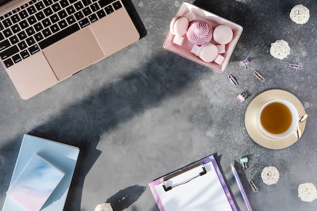 Postura plana de artigos de papelaria de escritório com uma xícara de chá com marshmallow e laptop em um fundo cinza com sombras. Foto Premium
