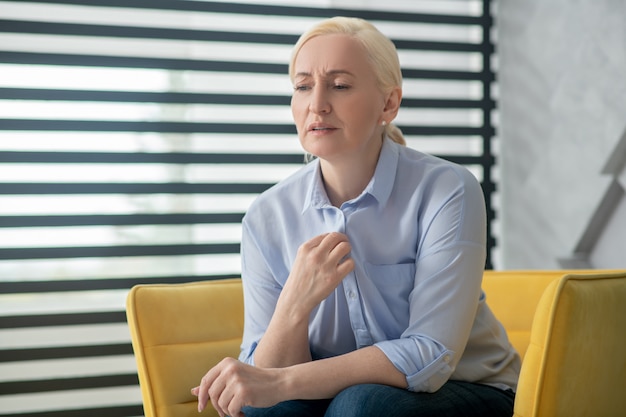 Reclamações, saúde. triste mulher adulta com cabelos loiros, sentado em uma cadeira perto da janela, falando na testa franzindo a testa. Foto Premium