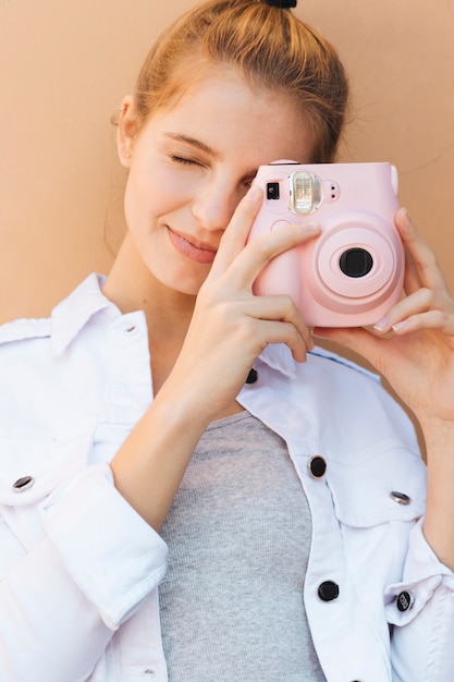 Retrato, de, um, mulher jovem, clicando, quadro, com, cor-de-rosa, câmera instantânea, contra, bege, fundo Foto gratuita