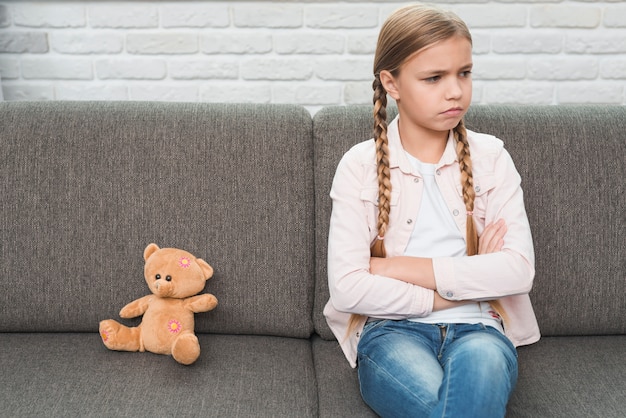 Retrato, de, um, triste, menina, com, braços cruzados, sentando, perto, a, urso teddy, ligado, cinzento, sofá Foto gratuita