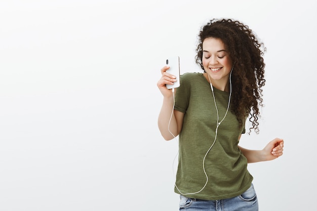 Retrato de uma menina bonita e elegante despreocupada com cabelos cacheados, dançando com os olhos fechados e um sorriso largo enquanto segura o smartphone e ouve música nos fones de ouvido Foto gratuita