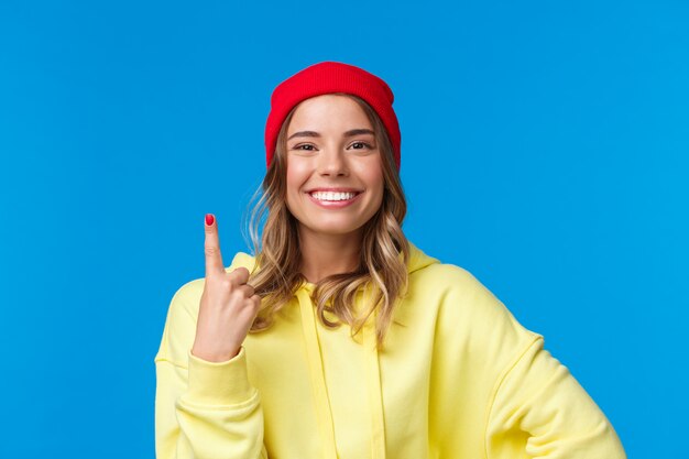 Retrato do close-up da fêmea europeia otimista bonito no gorro vermelho e capuz amarelo, mostrando o número um, ordem mesa única pessoa no café, sorrindo feliz, em pé | Foto Premium