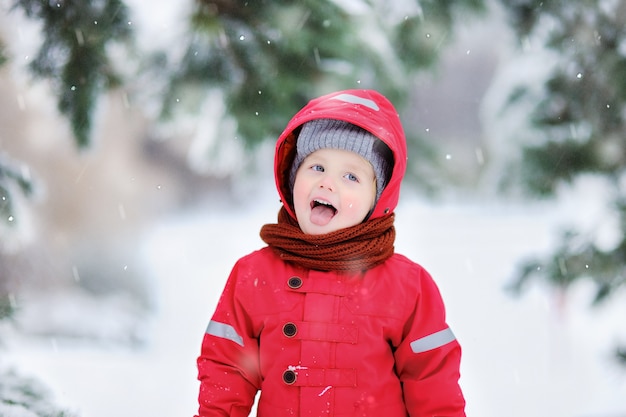Retrato do rapaz pequeno engraçado na roupa vermelha do inverno que tem o divertimento na queda de neve. lazer ativo ao ar livre com crianças no inverno. garoto com chapéu quente, luvas de mão e cachecol Foto Premium