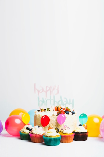 Saboroso bolo fresco com frutas e feliz aniversário título perto conjunto de bolinhos e balões Foto gratuita
