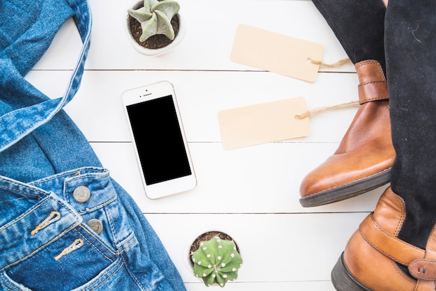 Smartphone perto de pano de brim, botas altas com etiquetas e cacto Foto gratuita