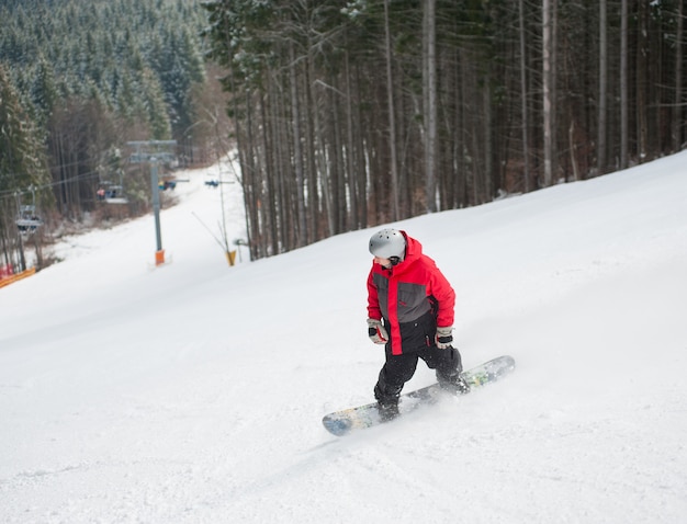 Snowboarder masculino desliza da montanha em dia de inverno, com vista para a encosta nevada e floresta em um resort de inverno Foto Premium