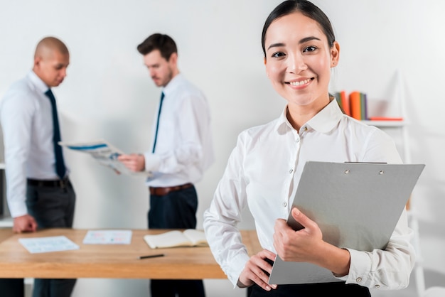 Sorrindo, jovem, executiva, segurando clipboard, em, mão, com, dois, homem negócios, trabalhando, em, fundo Foto gratuita