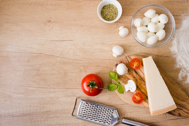 Tigela de vidro de queijo mussarela; tomates; manjericão e queijo bloco com ralador na mesa Foto gratuita