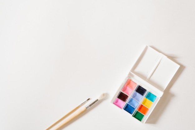 Tintas aquarela de cores diferentes nas valas em uma caixa branca e pincéis com alças de madeira sobre um fundo branco. copie o espaço. desenho e master classes para escola de arte Foto Premium
