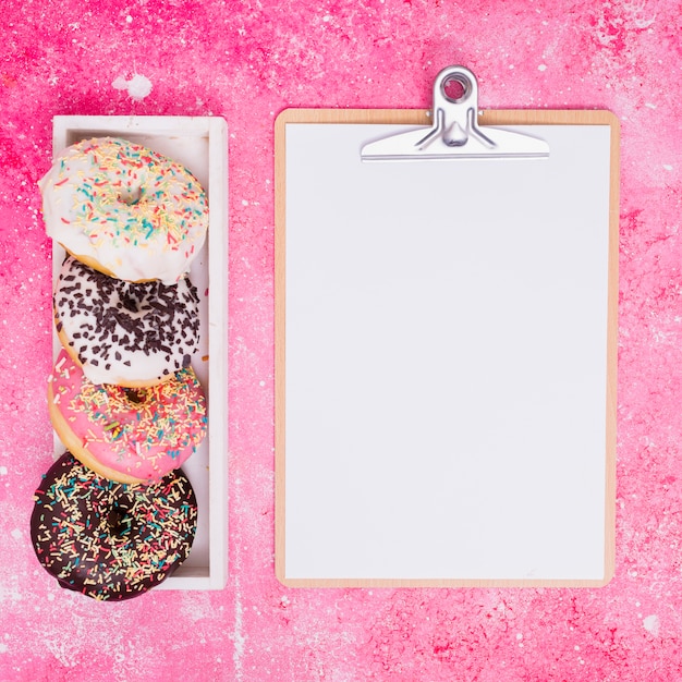 Tipo diferente de donuts em caixa retangular branca perto da prancheta com papel branco contra um fundo rosa Foto gratuita