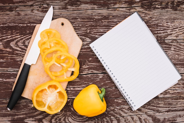 Todo e corte o pimentão amarelo na tábua de cortar com faca e espiral o bloco de notas na mesa de madeira Foto gratuita