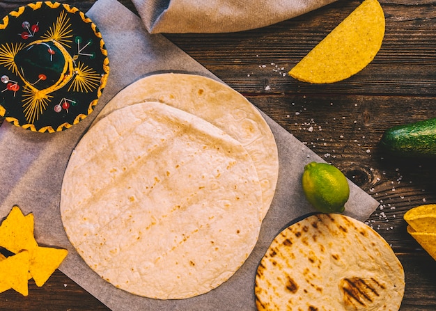 Tortilha mexicana de trigo; saborosos nachos e limões na mesa de madeira com chapéu mexicano Foto gratuita