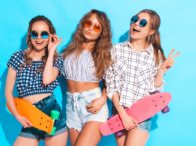 Três jovens elegantes sorridentes meninas bonitas com skates centavo colorido. mulheres em roupas de verão, posando em óculos de sol. modelos positivos se divertindo Foto gratuita