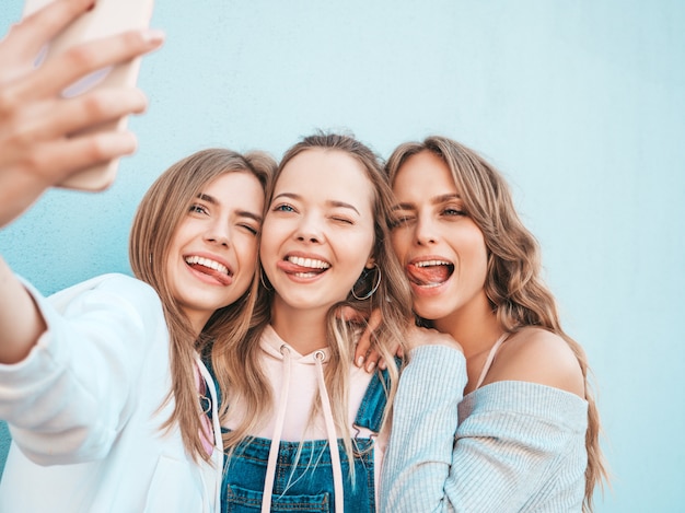 Três jovens sorrindo hipster mulheres em roupas de verão. meninas tirando fotos de auto-retrato de selfie em smartphone. modelos posando na rua perto da parede. feminino mostrando emoções de rosto positivo Foto gratuita