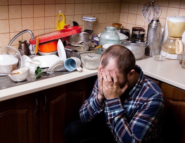 Premium Photo | A young bearded man sits on the floor and is shocked by the amount of dirty dishes in the kitchen sink to be washed.