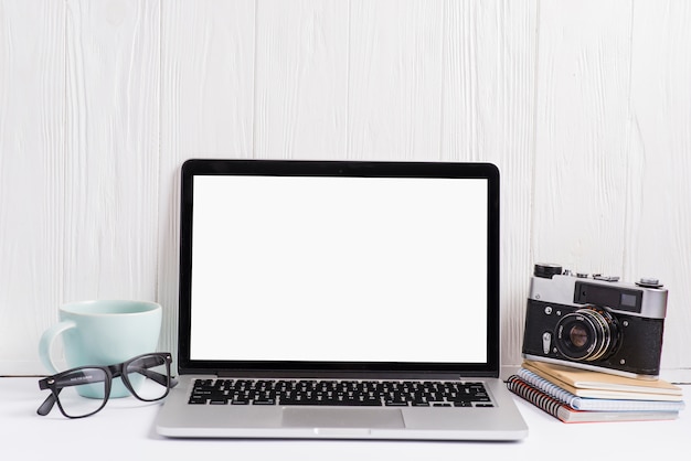 Um laptop aberto exibindo tela branca em branco com copo; óculos; câmera e notebook na mesa branca contra a parede de madeira Foto gratuita