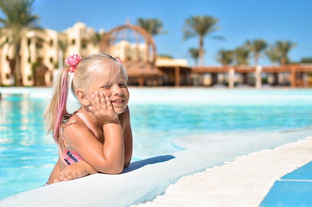 Uma menina de maiô senta-se na água azul de uma piscina em um hotel com palmeiras Foto Premium