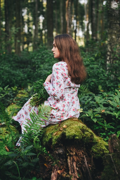 Uma mulher bonita em um vestido floral está sentada com um buquê de samambaia na floresta. Foto Premium