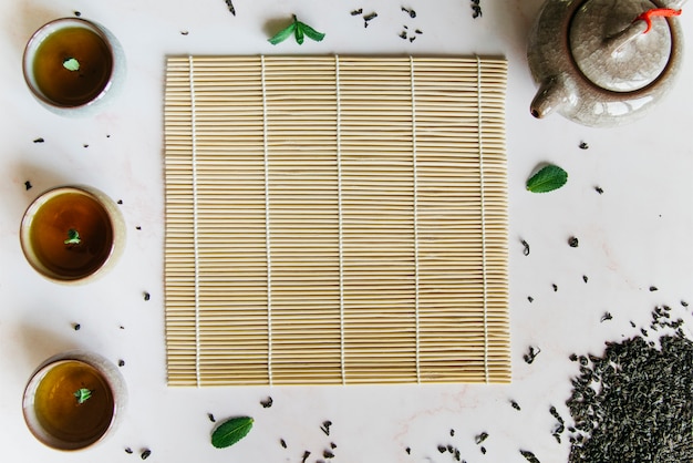 Uma visão elevada de xícaras de ervas com placemat; bule e folhas secas no pano de fundo branco Foto gratuita