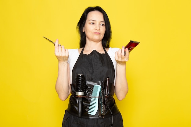 Uma vista frontal lindo cabeleireiro feminino na capa branca de camiseta preta com escovas com cabelos lavados, segurando uma tesoura e uma máquina posando no cabeleireiro estilista de fundo amarelo Foto gratuita