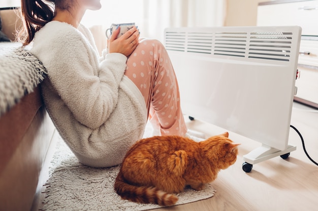 Usando aquecedor em casa no inverno. mulher aquecendo e bebendo chá com gato. estação de aquecimento. Foto Premium