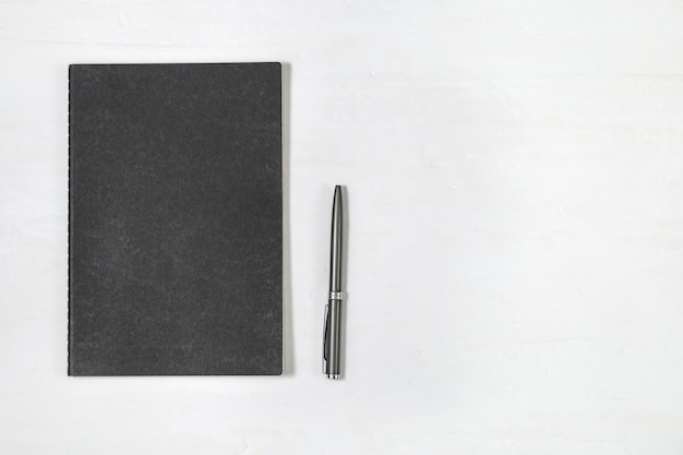 Vista superior do caderno fechado da tampa preta com a pena brilhante no fundo branco da mesa. mock up copybook. mínima mesa de escritório com artigos de papelaria. Foto Premium