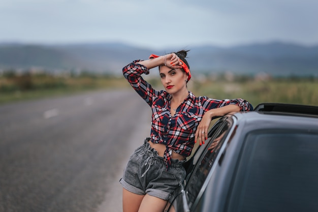 Vista traseira do jovem em shorts jeans curto cinza está consertando o carro. em shorts perto de um carro preto com o capô aberto. problemas com o carro na viagem. morena reparando o motor Foto Premium