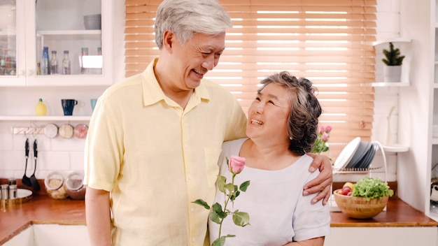 Asiatische ältere paare, die dem glücklichen lächeln und halten der blume und schauen zur kamera sich fühlen, während zu hause in der küche sich entspannt. lebensstil-ältere familie genießen konzept der zeit zu hause. porträt, blick in die kamera. Kostenlose Fotos