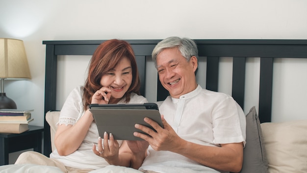 Asiatische ältere paare unter verwendung der tablette zu hause. asiatische ältere chinesische großeltern, ehemann und frau, die nach glücklich sind, wachen auf und passen film auf, auf bett im konzept des schlafzimmers zu hause morgens zu liegen. Kostenlose Fotos