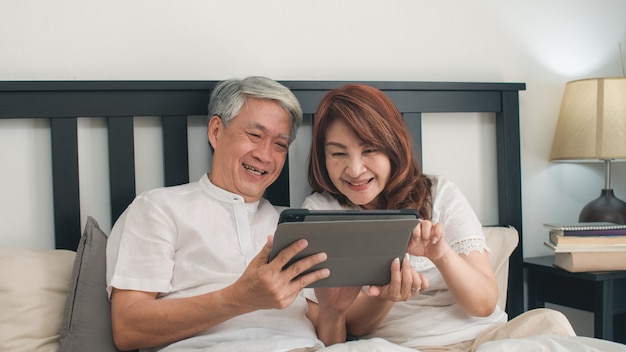 Asiatische ältere paare unter verwendung der tablette zu hause. asiatische ältere chinesische großeltern, ehemann und frau, die nach glücklich sind, wachen auf und passen film auf, auf bett im konzept des schlafzimmers zu hause morgens zu liegen. Kostenlose Fotos