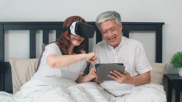 Asiatische ältere paare unter verwendung der tablette zu hause. asiatische ältere chinesische großeltern, glücklichem spaß glaubend und vr, das zusammen spiele beim auf bett im konzept des schlafzimmers zu hause liegen spielt. Kostenlose Fotos