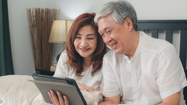 Asiatische ältere paare unter verwendung der tablette zu hause. asiatische ältere chinesische großeltern, videoanruf, der mit familienenkelkind spricht, scherzt beim auf bett im konzept des schlafzimmers zu hause morgens liegen. Kostenlose Fotos