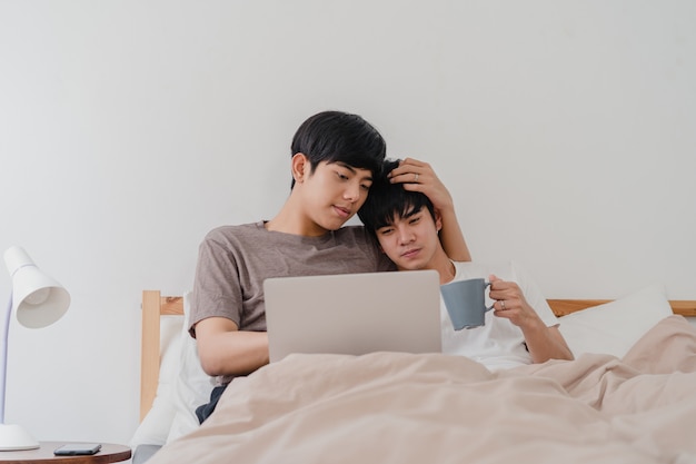 Asiatische homosexuelle mannpaare unter verwendung des computerlaptops und des trinkenden kaffees am modernen haus. das junge männliche glückliche asien-liebhaber entspannen sich rest zusammen, nachdem sie aufwachen und den film aufpassen, der auf bett im schlafzimmer am haus am morgen liegt Kostenlose Fotos