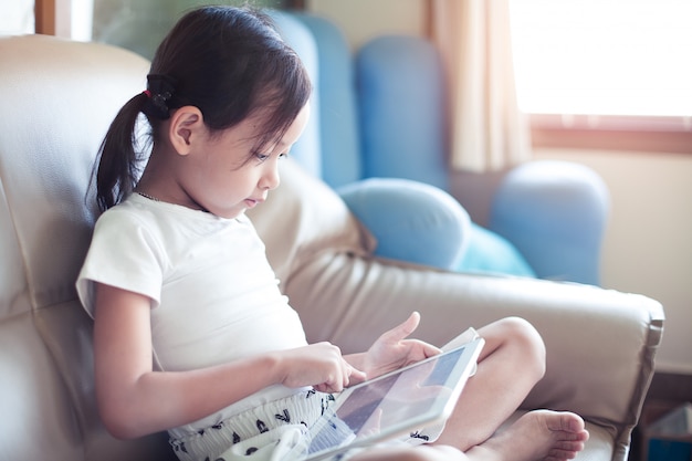 Asiatisches lächeln des kleinen mädchens, das auf sofa unter verwendung des schauens der digitalen tablettenauflage im wohnzimmer zu hause sitzt. Premium Fotos